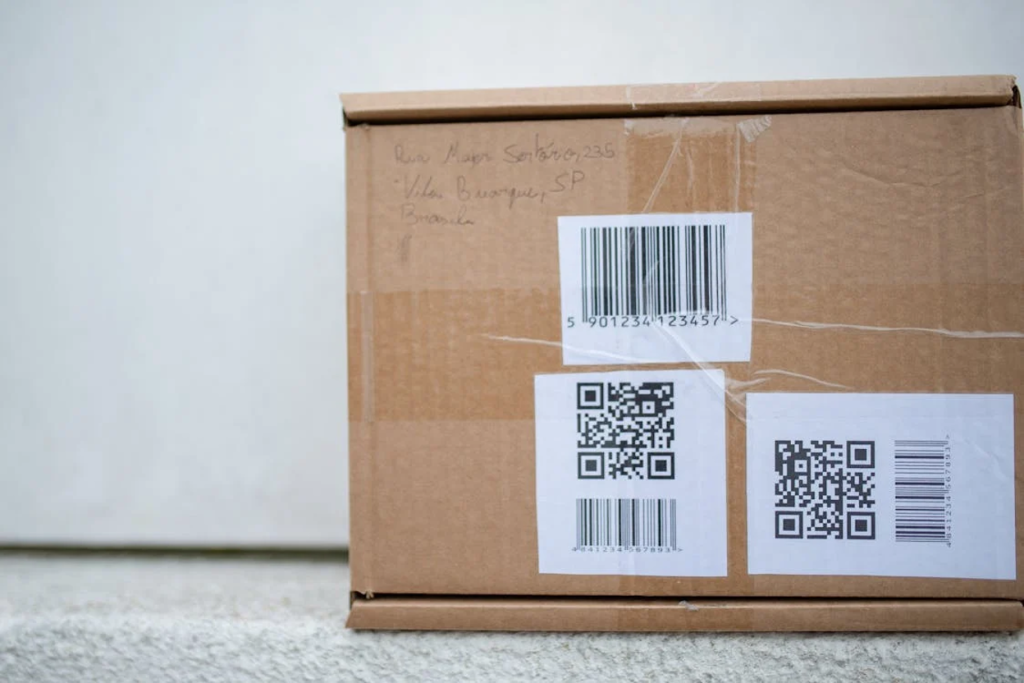Brown cardboard shipping box with handwritten address, barcodes, and QR code labels