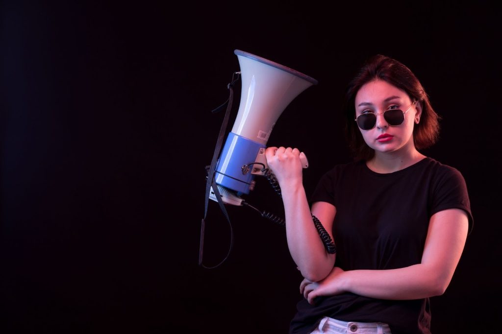 A young woman in a black t-shirt holding a megaphone under dramatic blue and pink lighting against a dark background.