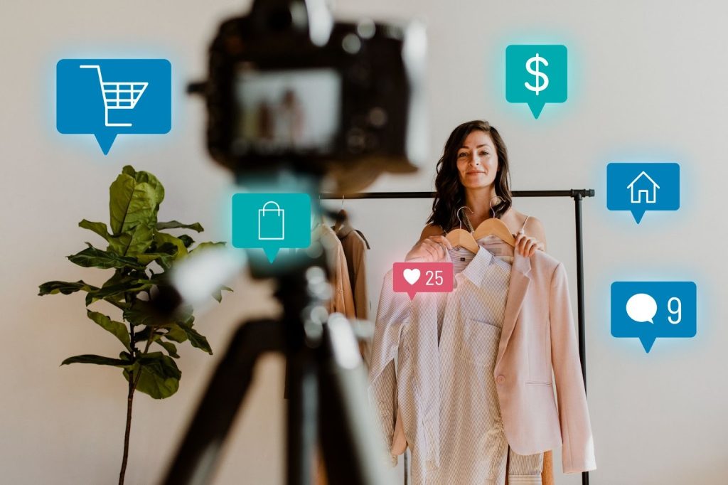 A woman holding clothing hangers in front of a camera on a tripod, surrounded by floating e-commerce and social media engagement icons.