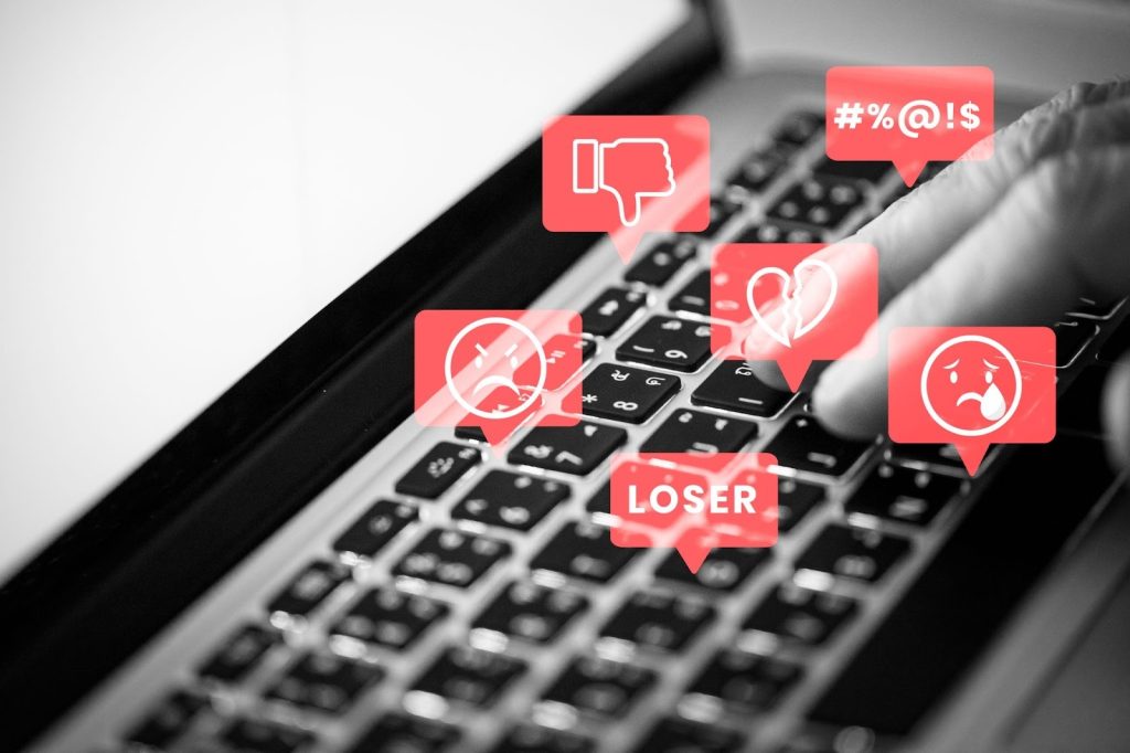Close-up of a laptop keyboard with floating red icons showing dislike, broken heart, angry emoji, and the word loser.