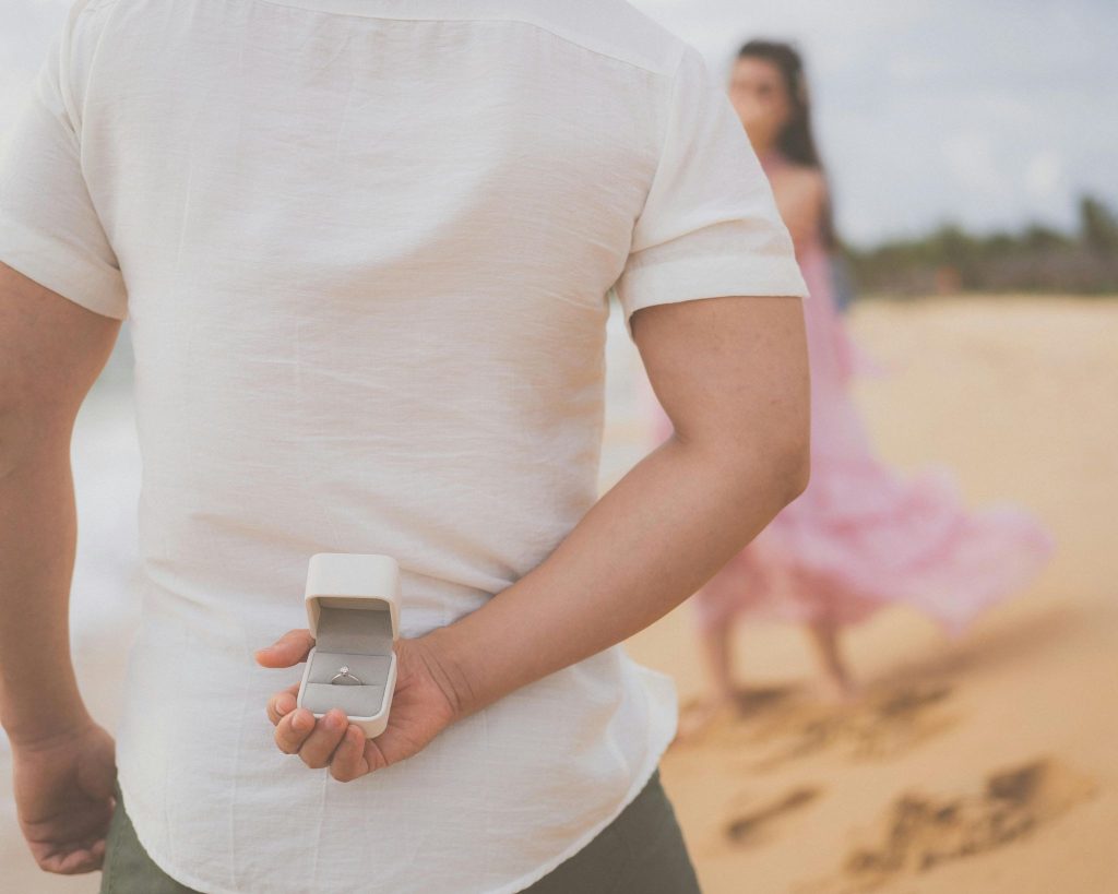 Man hiding open ring box behind back as woman in pink dress approaches on beach for proposal day