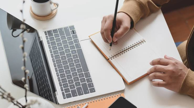 Person writing podcast show notes in a notebook beside an open laptop and a cup of coffee.