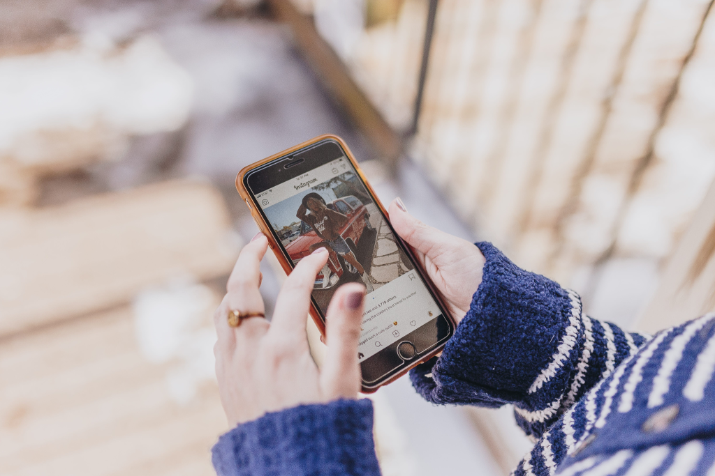 Hands holding a phone scrolling Instagram, tapping a photo on the screen.
