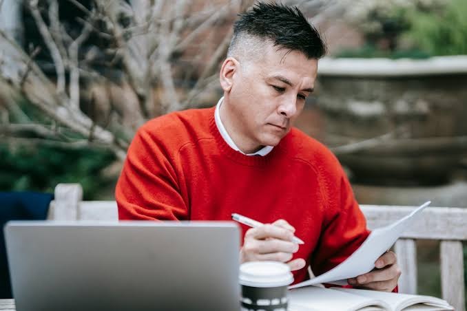 Man in red sweater reviewing printed report beside a laptop and coffee cup outdoors.